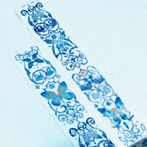 May include: Two rolls of blue and white patterned washi tape with a butterfly design. The tape is on a white background.