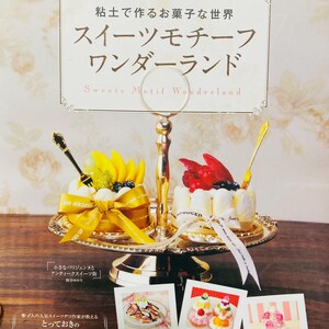 May include: A two-tiered gold metal serving stand holding two miniature cakes decorated with fruit, each wrapped in a ribbon. The top sign reads "粘土で作るお菓子な世界 スイーツモチーフワンダーランド". Below are three small cards showing various miniature dessert crafts.
