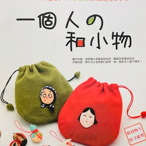 May include: A book cover with the title "One Person and Small Things" in Japanese characters. The cover features two fabric drawstring bags, one green and one red, with embroidered faces. The bags are decorated with small charms, including a bird, a flower, and a pair of traditional Japanese sandals.