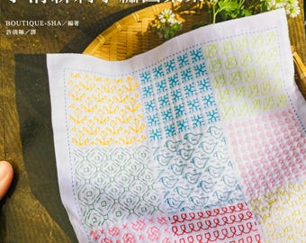 May include: A book cover featuring a white cloth with colorful embroidered patterns. The cloth is divided into squares with various designs in yellow, blue, green, pink, and red. Japanese text is visible at the top and bottom of the image.