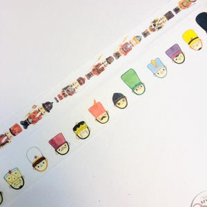 Könnte beinhalten: Dekoratives Washi-Tape mit einem sich wiederholenden Muster von Cartoon-Soldaten in roten, schwarzen und goldenen Uniformen. Darunter zeigt ein zweiter Streifen Cartoon-Gesichter mit verschiedenen Hutdesigns in Gelb, Grün und Lila. Das Tape befindet sich auf weißem Hintergrund.