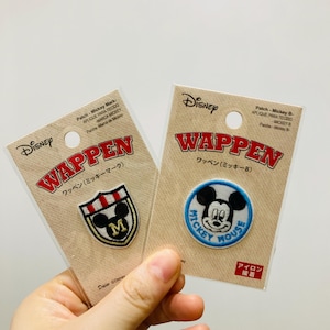 May include: Two Disney Mickey Mouse patches on cardboard backing. One patch is a shield with a black and white design and the letter "M". The other is a circular patch with Mickey Mouse and the words "Mickey Mouse".