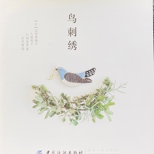 May include: A blue and gray embroidered bird with black accents, holding a green twig in its beak. The bird is perched on a branch of a small evergreen tree. The text "鸟刺绣" is written in black above the bird.