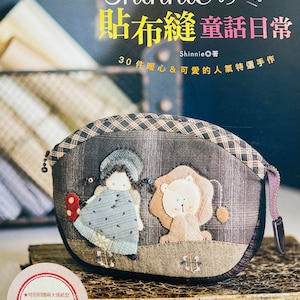 Könnte beinhalten: Buchcover mit einer handgefertigten Stofftasche mit einem skurrilen Design. Die Tasche ist mit applizierten Figuren eines Mädchens und eines Löwen verziert. Der Buchtitel lautet "Shinnie's Applique Fairy Tale Daily Life" mit japanischem Text und Illustrationen.