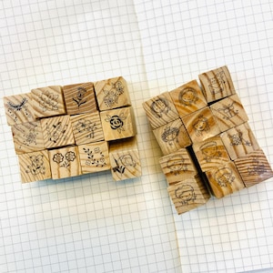 May include: Two sets of wooden stamps with various designs. One set features floral and botanical illustrations, including flowers, leaves, and a butterfly. The other set showcases illustrations of children and gift boxes. The stamps are arranged on a grid-lined paper.