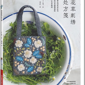 May include: A book titled "Flower and Grass Embroidery" featuring a gray tote bag with floral embroidery. The bag has white flowers, blue leaves, and brown accents. The book is surrounded by greenery in a white enamel bowl.