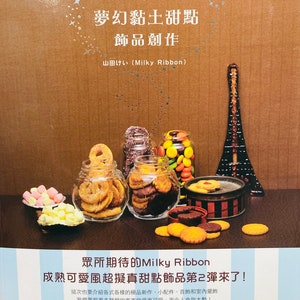 May include: A book cover with a brown background and a title that reads "Dreamy Clay Desserts Jewelry Creation" in Japanese. The cover features a miniature Eiffel Tower, jars of colorful candies, and a variety of miniature desserts.