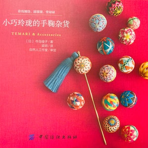 May include: A book cover with the title "Small and Exquisite Temari Accessories" in Japanese characters. The cover features a red background with a variety of colorful, embroidered temari balls, a blue tassel, and a gold needle.