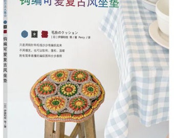 Puede incluir: Un libro titulado "钩编可爱复古风坐垫" (Hooked Cute Retro Style Cushion) con un cojín de ganchillo en tonos marrones, amarillos y verdes sobre un taburete de madera. La portada del libro presenta un mantel a cuadros y una taza y un platillo.