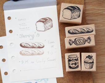 May include: A collection of wooden stamps featuring food illustrations, including bread, a candy, a jar of jam, and a cupcake. A notepad with bread sketches and text is held by a clipboard.