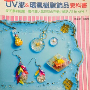 May include: A collection of handmade resin jewelry and accessories, including earrings, necklaces, and decorative pieces, displayed on a light blue tray. The jewelry features colorful designs and embedded elements. The image also shows a book with Japanese text.