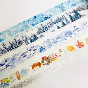 May include: A selection of washi tape rolls with winter-themed designs. The tapes display illustrations of snowflakes, snowy landscapes, and winter scenes in shades of blue and white. Some tapes have repeating snowflake patterns. The tape is measured in centimetres.