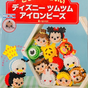 May include: A book cover with a red and white title that reads "ころんとかわいい ディズニー ツムツム アイロンビーズ"  The cover features a variety of colorful Disney Tsum Tsum characters made with beads. The book is part of the "レディブティックシリーズ" and is written by "セナパバ".