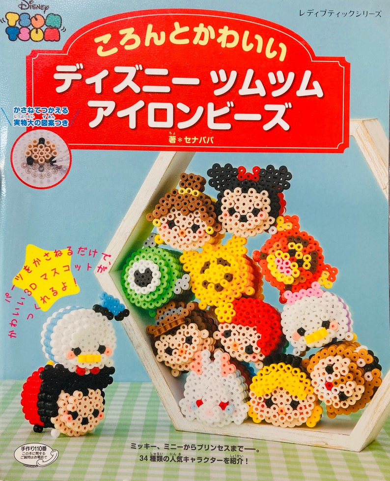 May include: A book cover with a red and white title that reads "ころんとかわいい ディズニー ツムツム アイロンビーズ"  The cover features a variety of colourful Disney Tsum Tsum characters made with beads. The book is part of the "レディブティックシリーズ" and is written by "セナパバ".