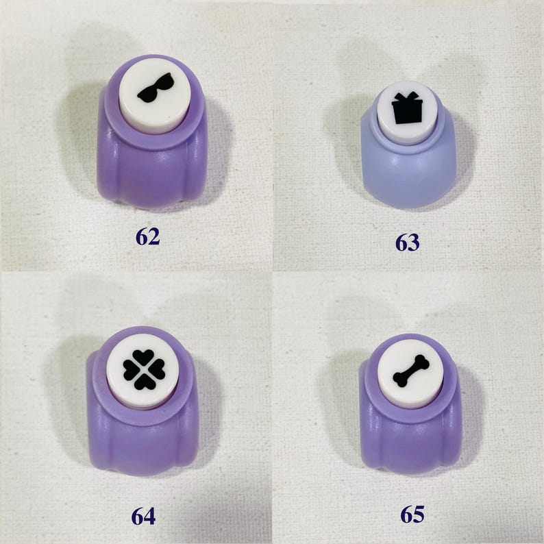 May include: Four purple craft punches labeled 62, 63, 64, and 65. Each punch features a white top with a black icon: sunglasses on 62, a gift box on 63, a four-leaf clover on 64, and a bone on 65. These tools are used for paper crafting.