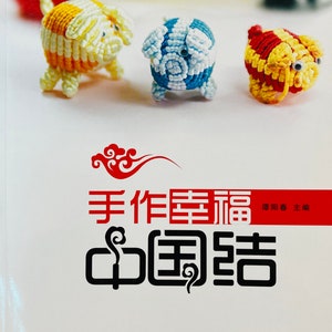 May include: A book cover with the title "Handmade Happiness Chinese Knots" in Chinese characters. The cover features three colourful Chinese knot pig figurines. The book is published by Liaoning Science and Technology Publishing House.