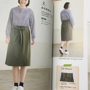 May include: A woman wearing a long-sleeved, blue and white striped shirt and an olive green skirt with a tie waist. The skirt falls to the mid-calf. The woman is wearing white sneakers and black socks. The image is from a fashion magazine.