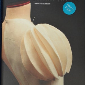 Puede incluir: Un libro titulado "Pattern Magic 2" de Tomoko Nakamichi, con un círculo azul que dice "Now in English". La portada presenta un primer plano de un maniquí de tela blanca con una manga plisada y fruncida.