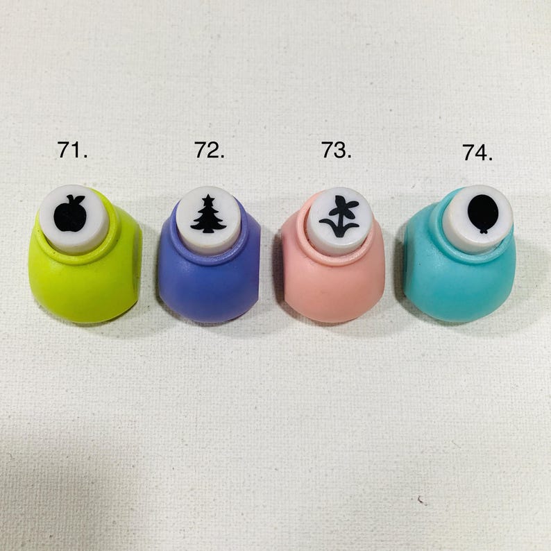 May include: Four craft paper punches in lime green, purple, light pink, and teal. Each punch features a white top with a black icon: 71. apple, 72. pine tree, 73. flower, and 74. balloon. These tools are designed for cutting shapes from paper.