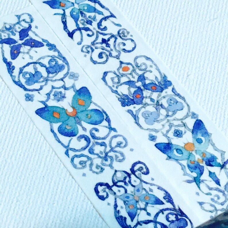 May include: Two rolls of blue and white patterned washi tape with a butterfly design. The tape is on a white background.