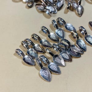 May include: A collection of small, silver-colored spoon-shaped pendants. Each pendant has a loop at the top and a textured bowl. The pendants are arranged on a light tan surface, with some scattered in the background.