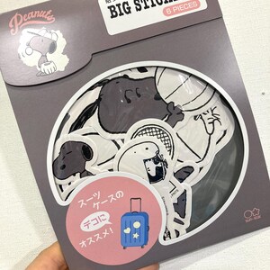 May include: A pack of Peanuts stickers featuring Snoopy and friends in black and white. The package, in grey and pink, is labelled "BIG STICKERS" and "6 PIECES". Japanese text is also visible on the packaging.