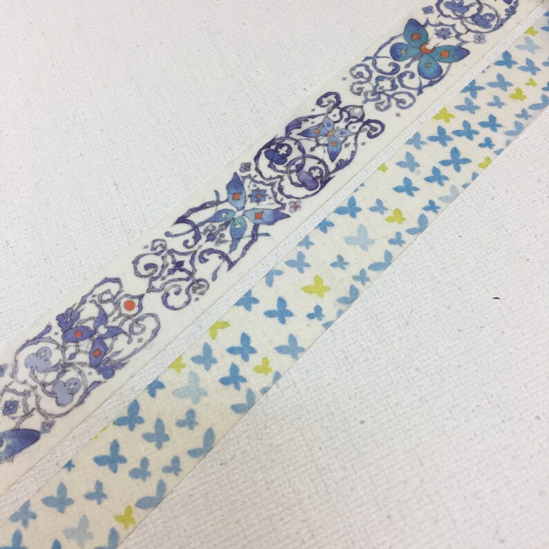 May include: Two rolls of washi tape with butterfly designs. One roll features a blue and white pattern with butterflies and floral accents. The other roll features a white background with blue and yellow butterflies.