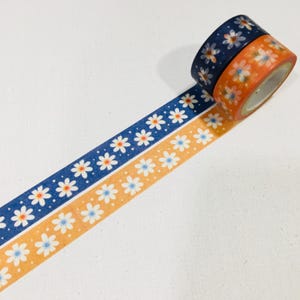 May include: Two rolls of adhesive washi tape, one dark blue and one orange, both featuring a repeating pattern of white daisy flowers with yellow centers. The tape is unrolled to show the floral design clearly against a plain white background.