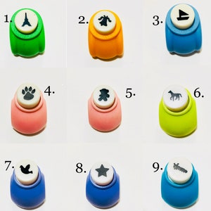 May include: A collection of nine colorful craft paper punches, each featuring a unique shape on top: an Eiffel Tower, a bell, a sailboat, a paw print, a teddy bear, a horse, a bird, a star, and a caterpillar, numbered one through nine.