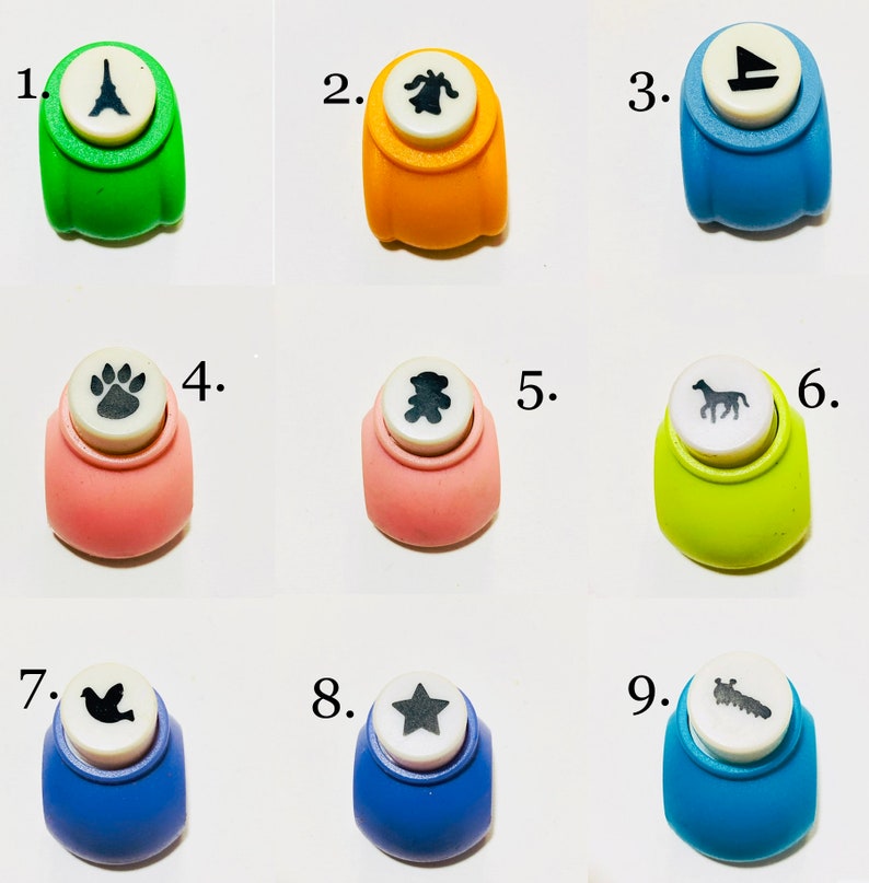May include: A collection of nine colorful craft paper punches, each featuring a unique shape on top: an Eiffel Tower, a bell, a sailboat, a paw print, a teddy bear, a horse, a bird, a star, and a caterpillar, numbered one through nine.