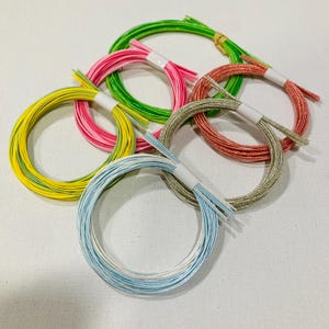 May include: Five circular coils of metallic cord in bright colors including lime green, hot pink, sunny yellow, copper, and light blue. Each coil is secured with a small white paper band, showcasing a shimmering, twisted texture throughout the strands.