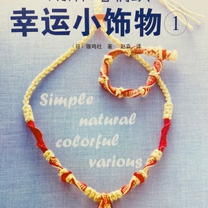 May include: A book cover with the title "Lucky Little Trinkets 1" in Japanese characters. The cover features a red and white braided cord necklace with a small, red and gold pendant. The book is about making simple, natural, colourful, and various trinkets.