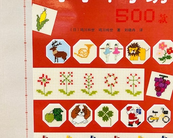 May include: A book cover with the title "Little Cross Stitch 500 Designs" in Japanese. The cover features a red background with a variety of colorful cross stitch designs, including a deer, a musical instrument, a flower, and a grape.