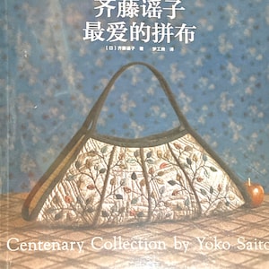 Op de afbeelding: Een boek getiteld "Centenary Collection by Yoko Saito" met een gestikte handtas met een bloemenpatroon. De boekomslag is blauw met witte stippen en de handtas is bruin met een lichtbruine achtergrond en een bloemenpatroon.