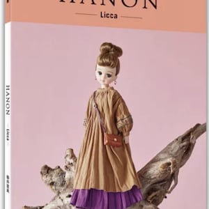 May include: A doll stands on a piece of driftwood in front of a pink background. The doll wears a brown dress with a purple hem and a small brown purse. The book cover reads "HANON - Licca".