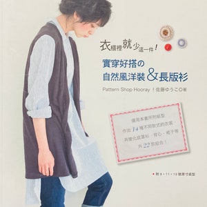 Op de afbeelding: Een boekomslag met een vrouw die een grijze vest draagt over een blauw-wit gestreept shirt en een blauwe jeans. De titel van het boek is "Real Wear Good Fit" en de tekst onder de titel is "Natuurlijke windjurk & Lange cardigan". De boekomslag bevat ook een klein wit label met de tekst "Gebruik het patroon van dit boek om 14 verschillende kledingstukken te maken, waaronder cardigans, vesten en rokken. Er zijn in totaal 22 combinaties!"