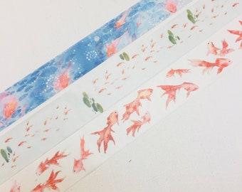 Könnte beinhalten: Drei Rollen Washi-Tape mit einem Aquarell-Design von Goldfischen, die im Wasser schwimmen. Die obere Rolle hat einen blauen Hintergrund mit weißen Punkten, die mittlere Rolle hat einen weißen Hintergrund mit grünen Blättern und die untere Rolle hat einen weißen Hintergrund ohne zusätzliches Design.