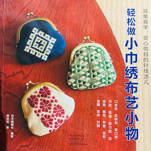 May include: A book cover with the title "Easy to Learn, Delightful Needlework:  Make Small Cloth Embroidery Crafts" in Japanese. The cover features three small, embroidered coin purses with different designs. The purses are on a wooden surface.