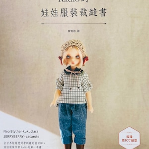 Può includere: Un libro intitolato "Radio's Doll Clothes Sewing Book" di Ji Eun Choi. La copertina presenta una bambola che indossa una camicia a quadri, jeans blu e un cappello all'uncinetto. Il libro include modelli per realizzare abiti per bambole.