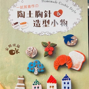 May include: A book cover with the title "Homemade Cookie Style" in a decorative font. The cover features a variety of colourful clay charms, including a house, a cat, a tree, a croissant, and a loaf of bread. The text "一試就會作の 陶土胸針 造型小物" is also on the cover.