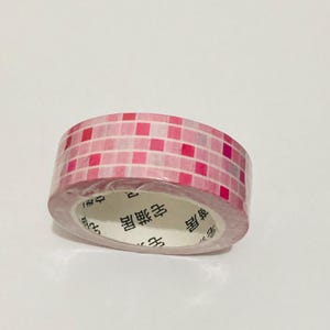 May include: A roll of decorative tape with a pink and red square pattern. The tape is wrapped around a white core with Japanese text. The tape is on a white background.