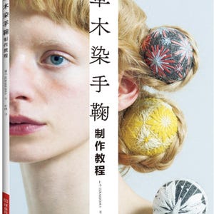 May include: A book titled "草木染手鞠制作教程" with Japanese text, featuring a woman's face and hair adorned with colorful Temari balls. The book's spine is visible, and the cover displays the title. The image has a soft, artistic aesthetic.