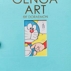 Könnte beinhalten: Hellblauer Buchumschlag mit dem Titel "THE GENGA ART OF DORAEMON" in goldfarbenen Buchstaben. Das Cover zeigt eine Illustration der Zeichentrickfigur Doraemon. Japanischer Text ist ebenfalls vorhanden.
