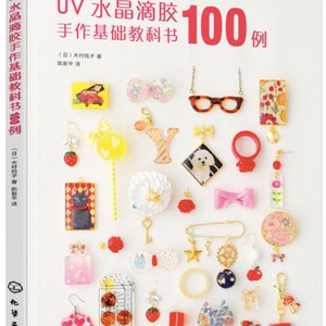 May include: A book cover with a white background and colorful resin crafts. The book title is "UV Resin Jewelry 100 Examples" in Japanese and English. The book cover features a variety of resin crafts, including earrings, necklaces, rings, and charms.