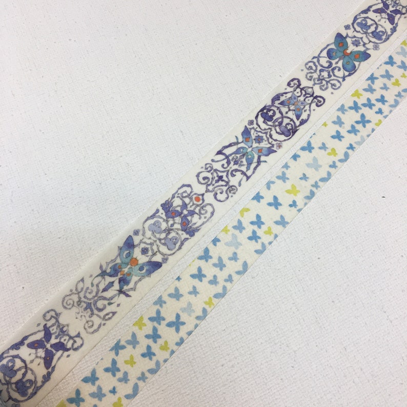 May include: Two rolls of washi tape with butterfly designs. One roll features a repeating pattern of blue butterflies on a white background. The other roll features a more intricate design with blue butterflies, swirls, and floral elements on a white background.