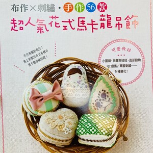 May include: A collection of five fabric and embroidery macaron-shaped coin purses in a wicker basket. The purses are decorated with various designs, including a heart, a flower, a checkered pattern, and a Russian nesting doll. The text on the page reads "macaron 可愛進化! 布作×刺繍・手作56款 超人氣花式馬卡龍吊飾"
