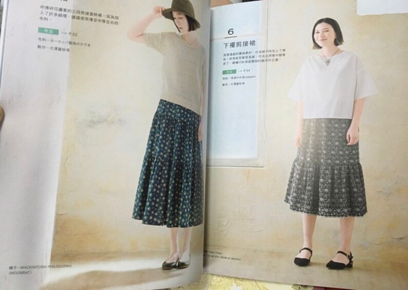 May include: Two women modeling outfits from a fashion magazine. One wears a beige sweater, a patterned skirt, and a hat. The other wears a white top and a patterned skirt. The magazine is open to a page with Japanese text.