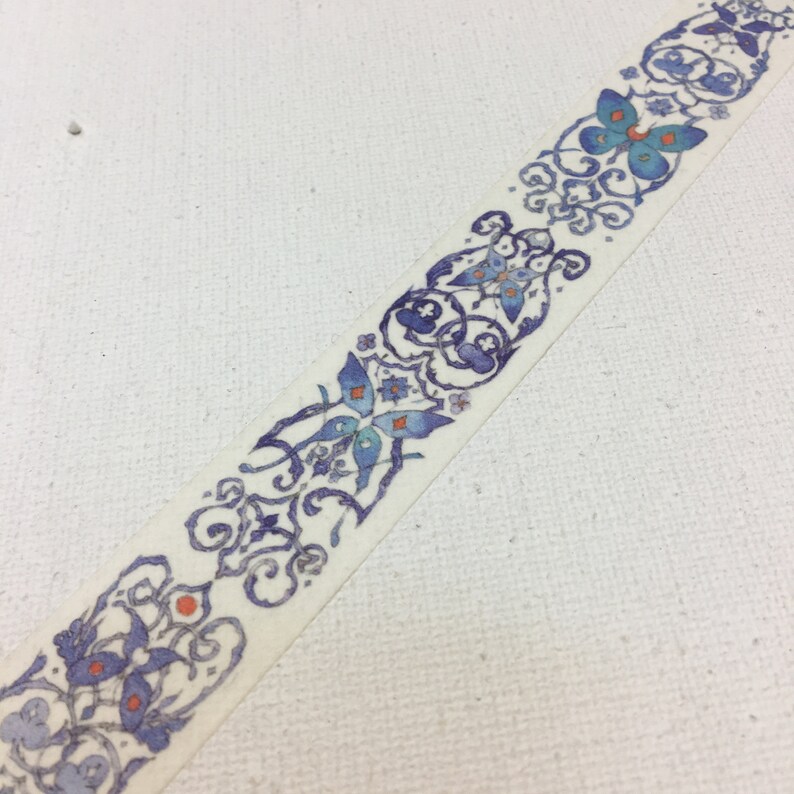 May include: A white washi tape with a repeating pattern of blue butterflies and floral designs. The butterflies have orange accents.
