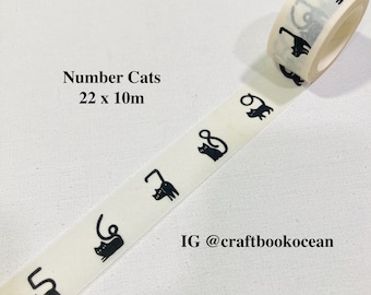 Puede incluir: Rollo de cinta washi decorativa con un patrón repetido de siluetas de gatos negros. La cinta está etiquetada como "Number Cats" y mide 22 mm x 10 m. También se ve el identificador de Instagram @craftbookocean.