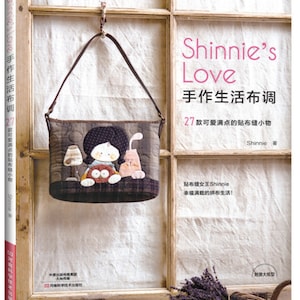 Puede incluir: Un libro titulado "Shinnie's Love" con texto japonés y una bolsa de tela decorativa colgada frente a una ventana. La bolsa presenta un diseño de niña de dibujos animados y conejo. La portada del libro es roja y blanca.